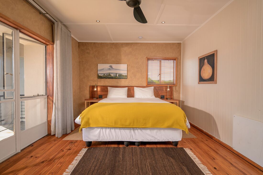 Un dormitorio con cama, ventilador de techo, ventana con persianas, un cuadro en la pared y puerta que da a un balcón.