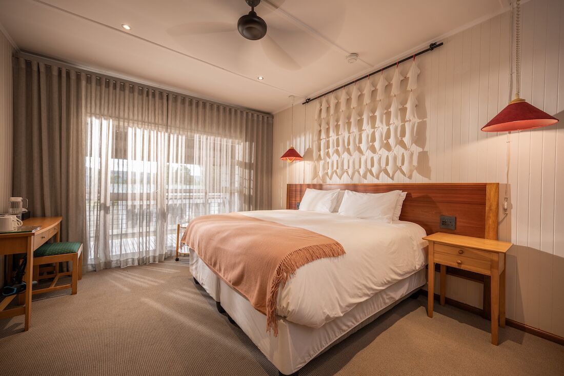 A hotel room with a large bed, a desk with a chair, a wooden nightstand, and a wall-mounted lamp.