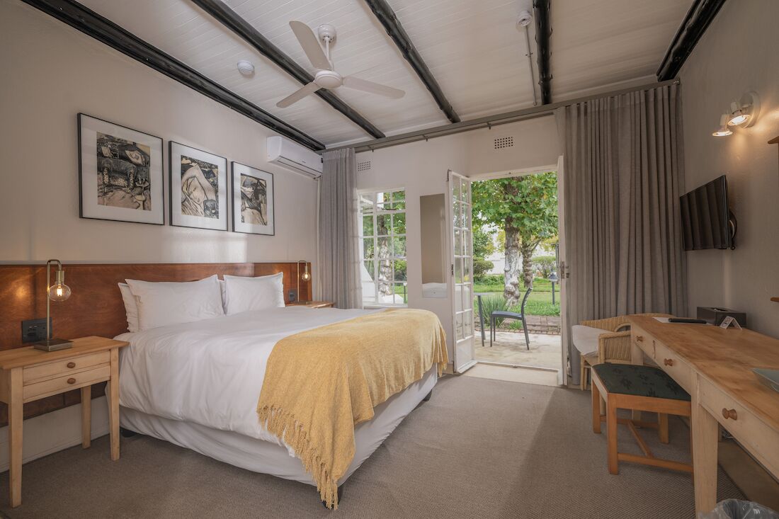 A hotel room with a bed, bedside tables, a desk, and a TV. There are framed pictures on the wall, a ceiling fan, and a view of the outdoors.