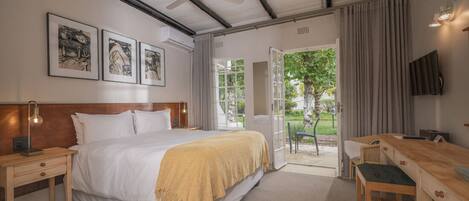 A hotel room with a bed, bedside tables, a desk, and a TV. There are framed pictures on the wall, a ceiling fan, and a view of the outdoors.