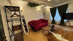 Una habitación con un sofá rojo, un espejo, suelo de madera, una planta colgante y un televisor sobre un trípode.