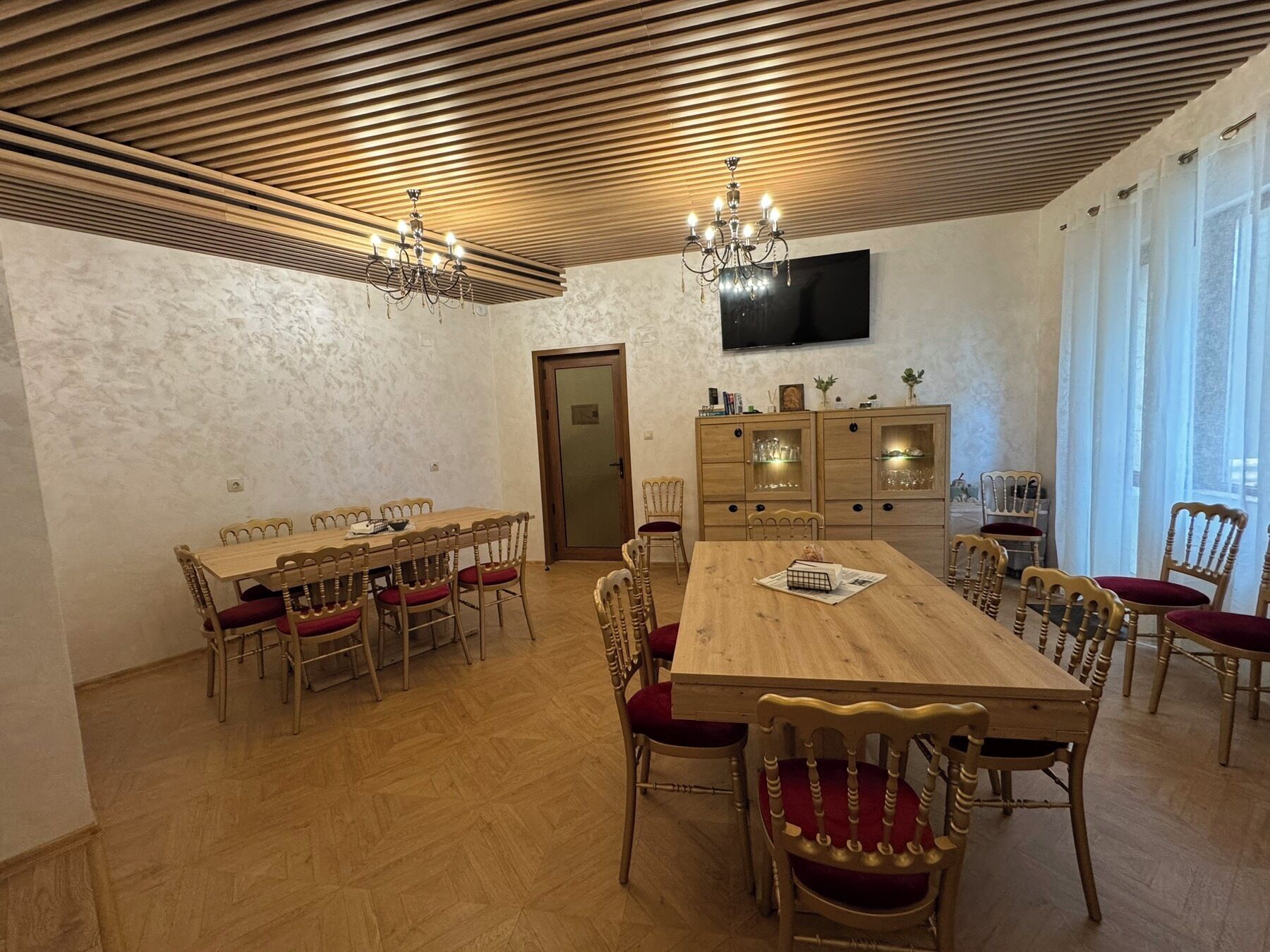 Un comedor con una mesa y sillas de madera, una araña, un televisor montado en la pared y una puerta.