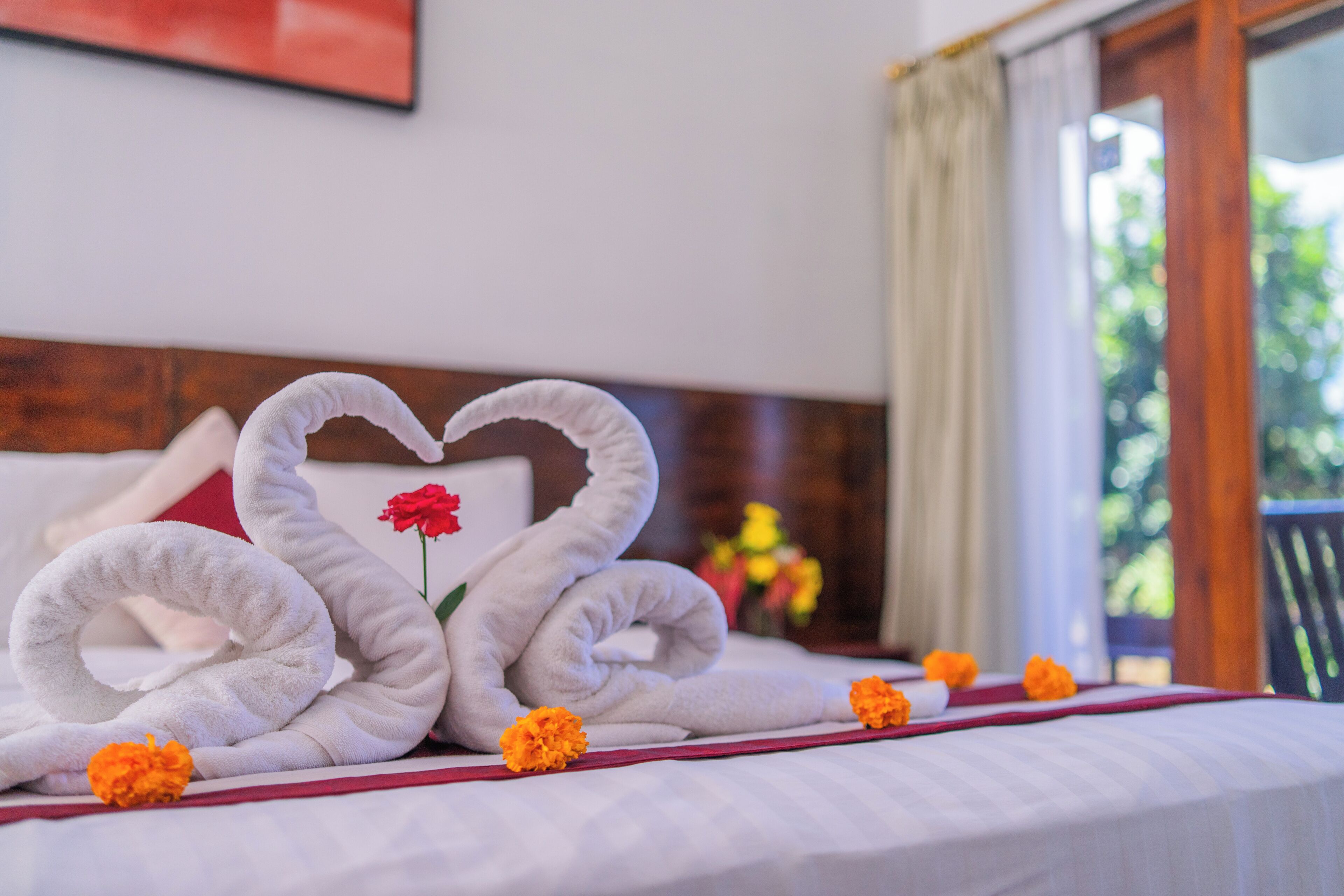 A bed with a heart-shaped towel arrangement and a single red flower.