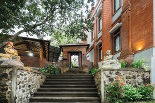 Una escalera conduce a la entrada de un edificio, flanqueada por estatuas y rodeada de follaje.