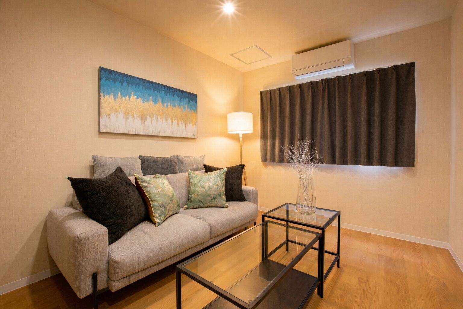 A living room with a sofa, a glass coffee table, a lamp, a painting on the wall, and an air conditioning unit.
