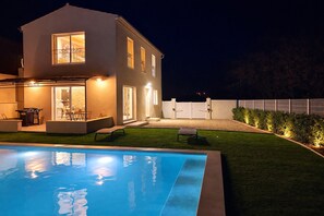 Una casa moderna de dos pisos con piscina, mobiliario exterior y una zona de patio bien iluminada.