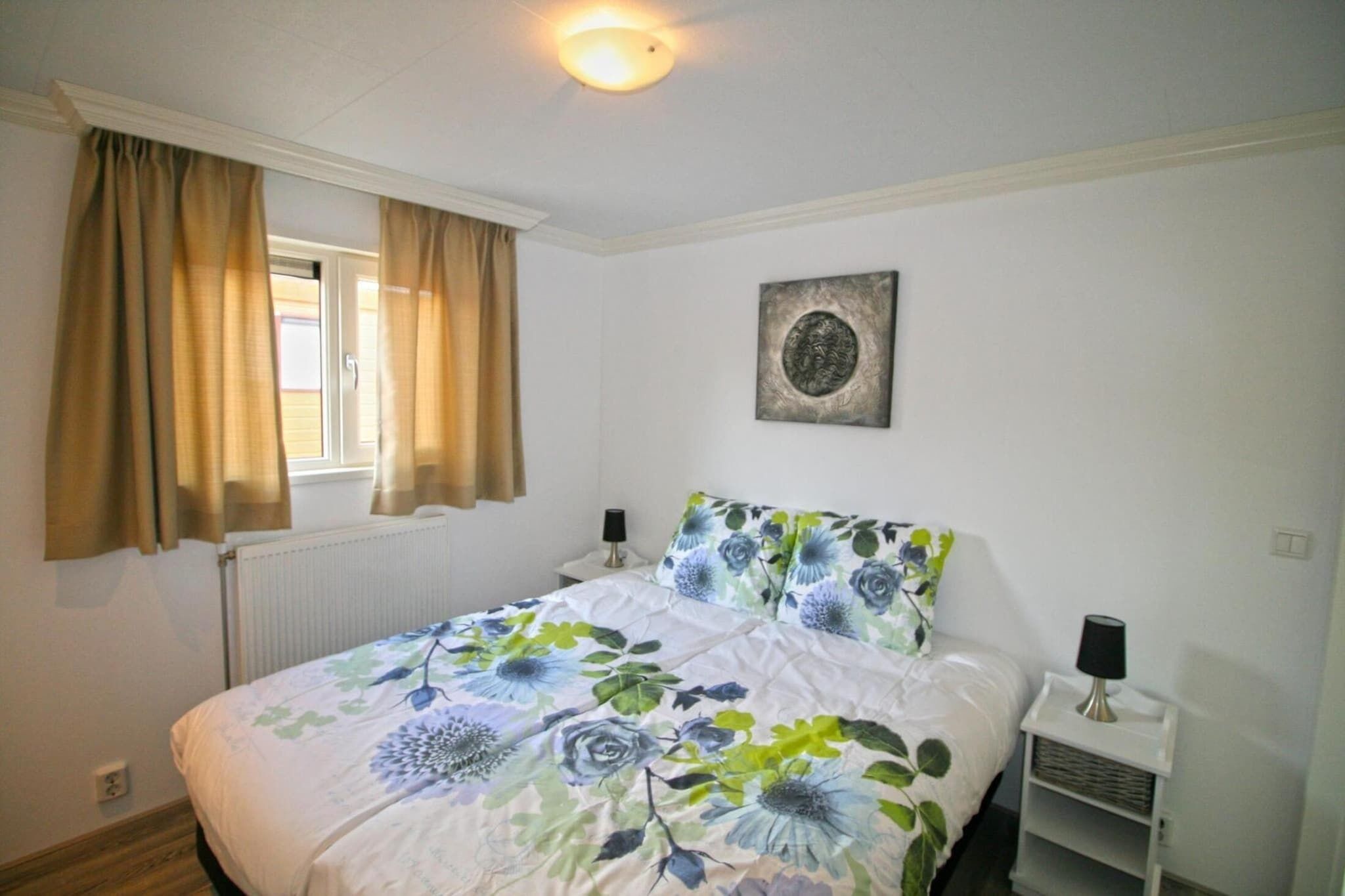 A bedroom with a bed, curtains, a wall-mounted lamp, a radiator, and a ceiling light fixture.