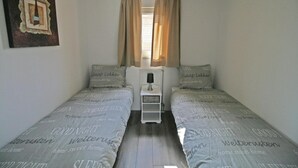 Un dormitorio pequeño y ordenado con dos camas individuales, una mesita de noche, una lámpara y una ventana con cortinas.