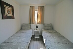 Un dormitorio pequeño y ordenado con dos camas individuales, una mesita de noche, una lámpara y una ventana con cortinas.