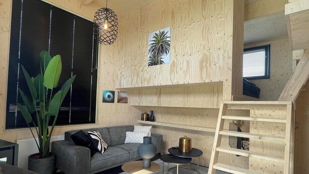 Ein modernes Interieur mit einem grauen Sofa, einem runden Couchtisch, Holzregalen und einem großen Fenster mit Jalousien.