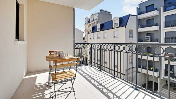 Ein Balkon mit Holztisch und -stühlen, einem Metallgeländer und Blick auf benachbarte Gebäude.