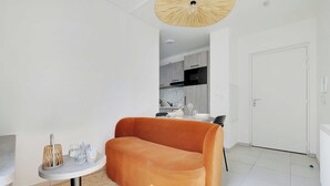 Ein modernes Wohnzimmer mit einer Couch, einem kleinen runden Tisch mit Lampe und einer Küche im Hintergrund.