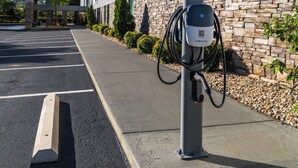 A parking area with a charging station for electric vehicles, a stone wall, and a building in the background. - Augusta West (Augusta)
