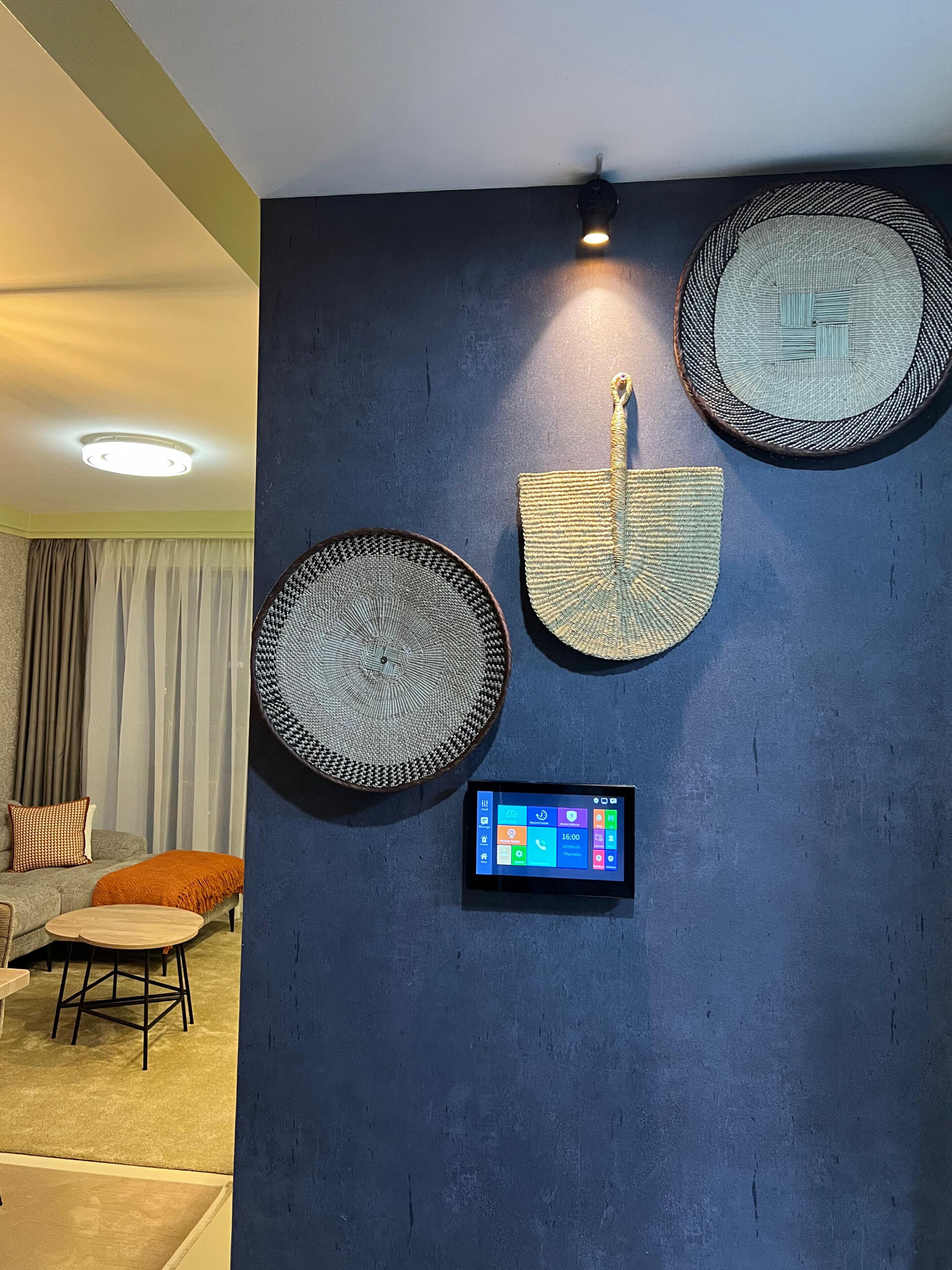Ein modernes Wohnzimmer mit dunkelblauer Akzentwand, einem runden Wandbehang aus Gewebe, einem Flachbildfernseher und einer gemütlichen Sitzecke mit Sofa und Hocker.