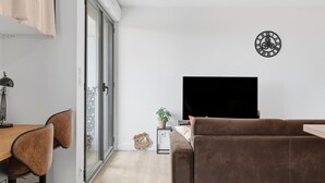 En moderne stue med et fladskærms-tv, en brun sofa, et spiseområde med stole, en lille plante og et dekorativt skjold på gulvet.