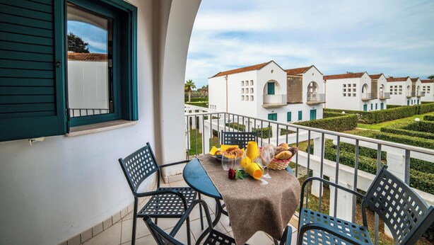 A balcony with a table set for two, a window with green shutters, and a view of white buildings with red roofs. - Blumare Apartment 3 Pool & Sea (Otranto)
