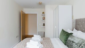 Een opgemaakt bed met witte lakens en een groen kussen, een nachtkastje met een lamp en een garderobe met een spiegel.