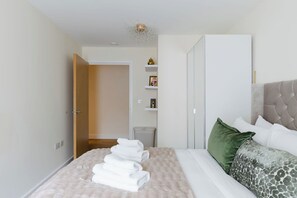 A neatly made bed with white linens and a green pillow, a bedside table with a lamp, and a wardrobe with a mirror. - Beautifully Presented 2BDR Flat in Canada Water (Greater London)