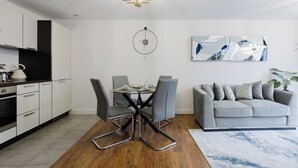 Een moderne keuken met witte kasten, een eethoek met een ronde tafel en stoelen, een grijze bank en een wandklok.