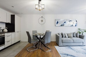 A modern kitchen with white cabinets, a dining area with a round table and chairs, a grey sofa, and a wall clock. - Beautifully Presented 2BDR Flat in Canada Water (Greater London)