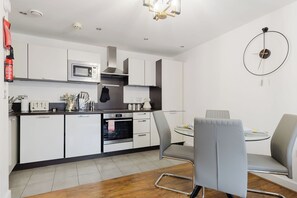 A modern kitchen with white cabinets, a microwave, a stove, and a dining area with a grey chair and table. - Beautifully Presented 2BDR Flat in Canada Water (Greater London)