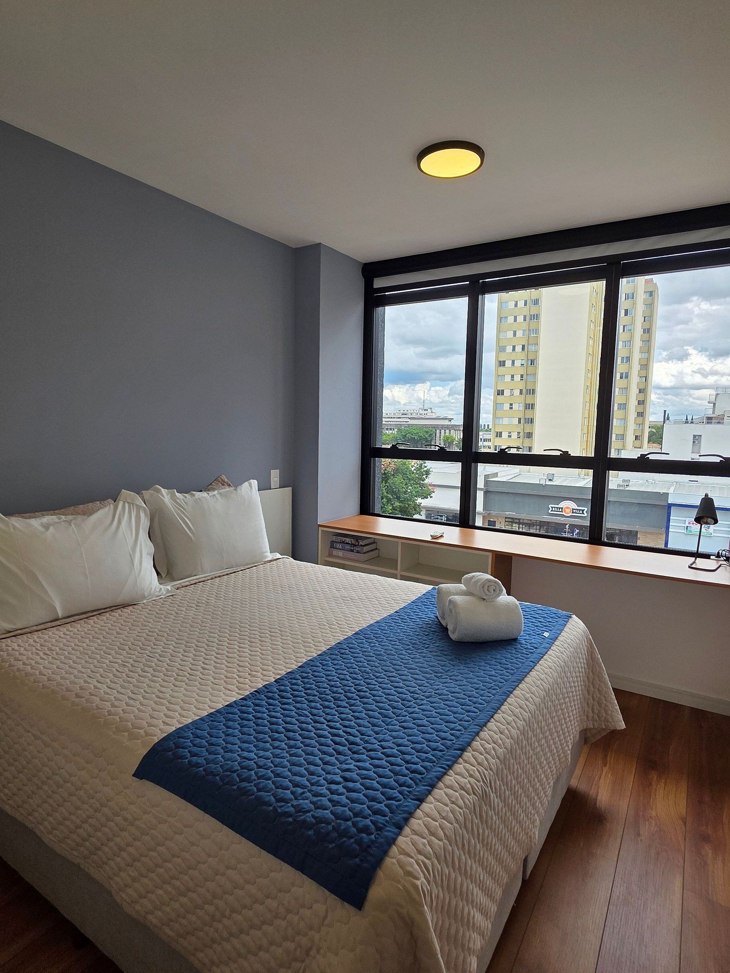 Une chambre avec un grand lit, une fenêtre donnant sur la ville, une table de chevet et un luminaire au plafond.