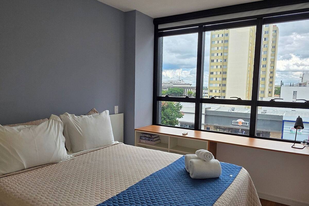 Quarto com uma cama grande, janela com vista para a cidade, mesa de cabeceira e luminária no teto.