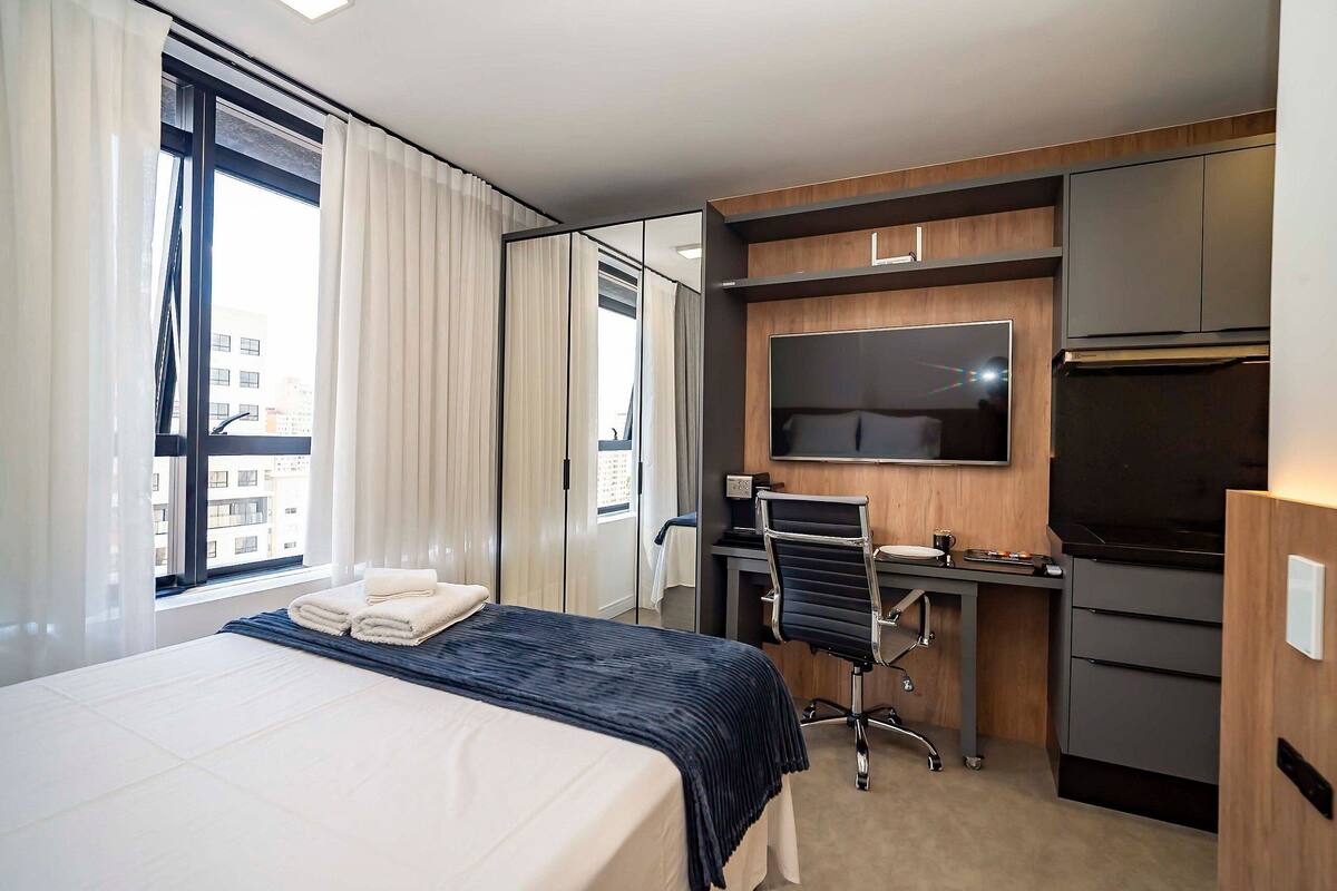 Quarto de hotel com cama, escrivaninha com cadeira, televisão de tela plana, frigorífico e janela com cortinas.