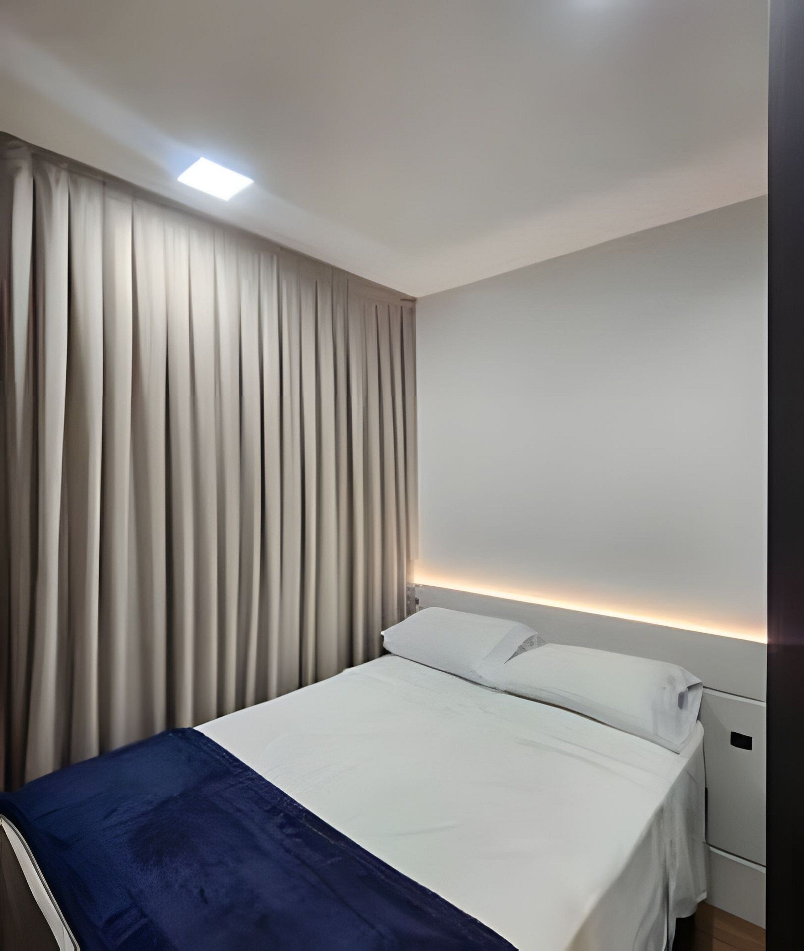 Quarto de hotel com cama, cortinas, luminária no teto e mesa de cabeceira.