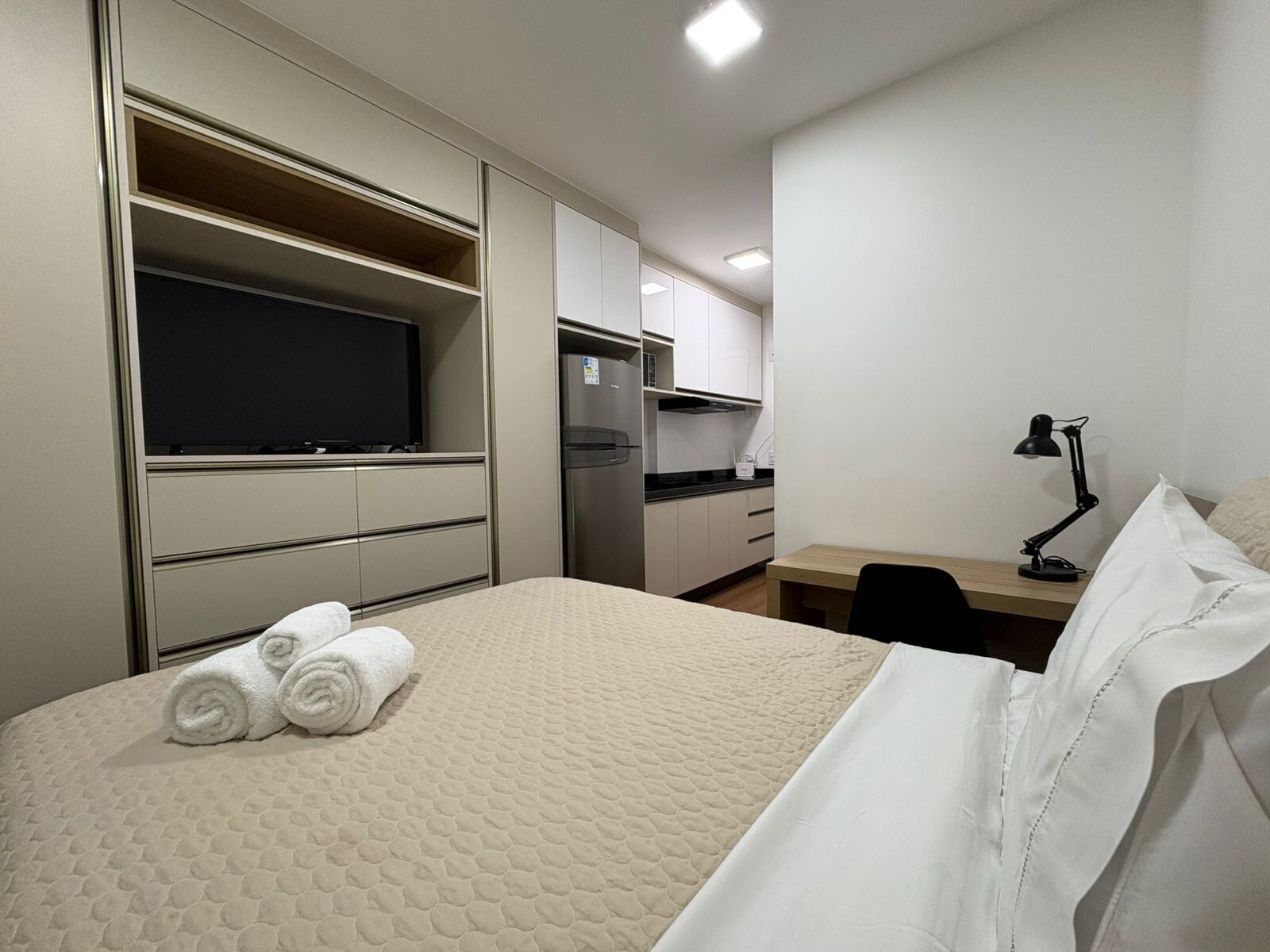 Quarto de hotel com cama, escrivaninha, televisão, frigorífico e candeeiro.