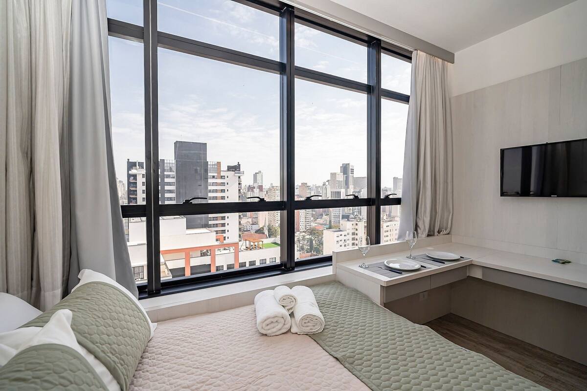 Quarto de hotel moderno com grandes janelas, uma cama, um espaço de estar pequeno, uma TV e vista para a paisagem urbana.