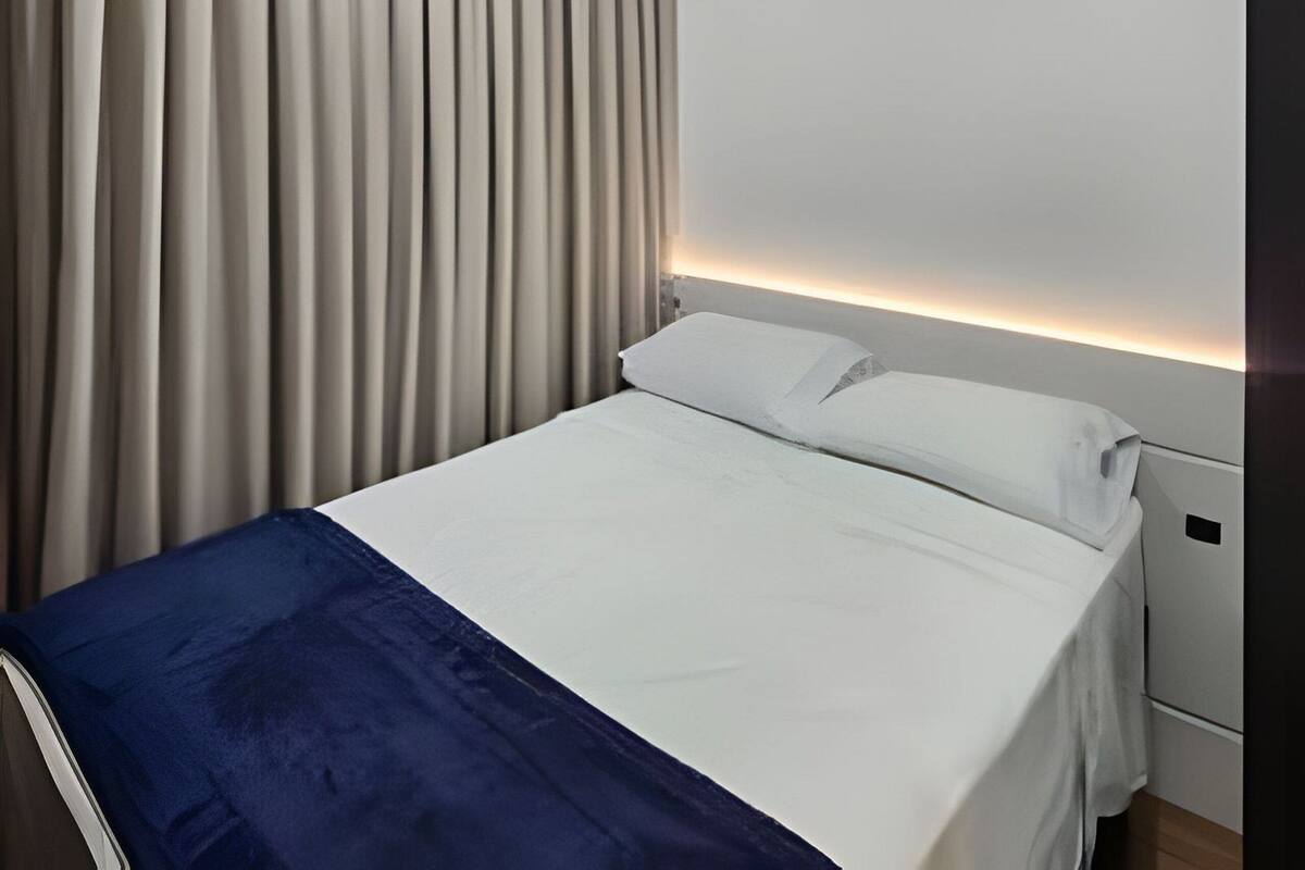 Quarto de hotel com cama, cortinas, luminária no teto e mesa de cabeceira.