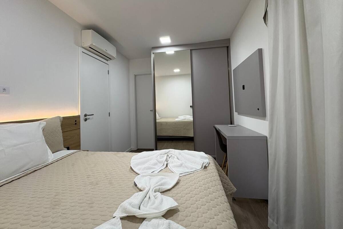 Quarto de hotel com cama, escrivaninha, aparelho de ar condicionado e espelho.