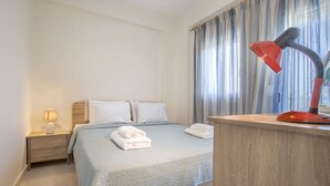 A hotel room with a bed, a nightstand with a lamp, a desk with a red lamp, and a window with curtains. - Sunrise Villa No 1 - Agios Nikolaos Halkidiki (Vourvourou)