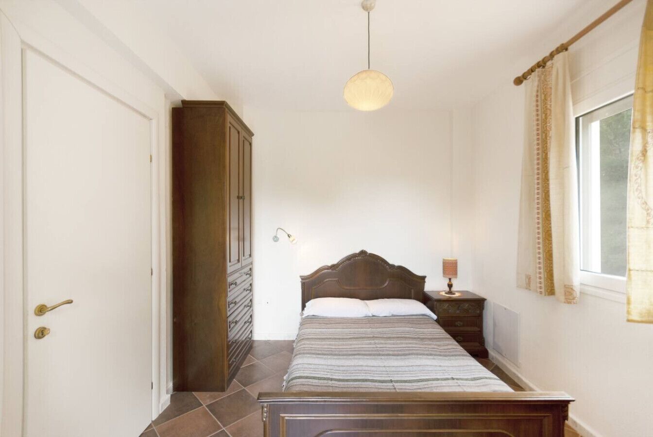 Ein Schlafzimmer mit Bett, Kleiderschrank, Nachttisch, Lampe und einem Fenster mit Vorhängen.