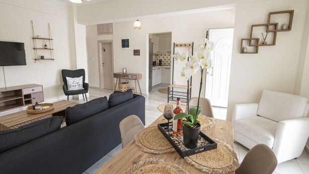 A modern living room with a sofa, dining table, and chairs, a TV, and a kitchen in the background.