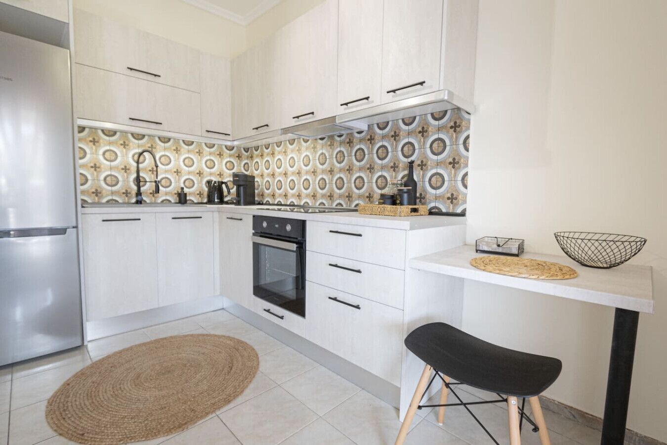 A compact kitchen with white cabinets, a stainless steel refrigerator, a black oven, and a small black stool.