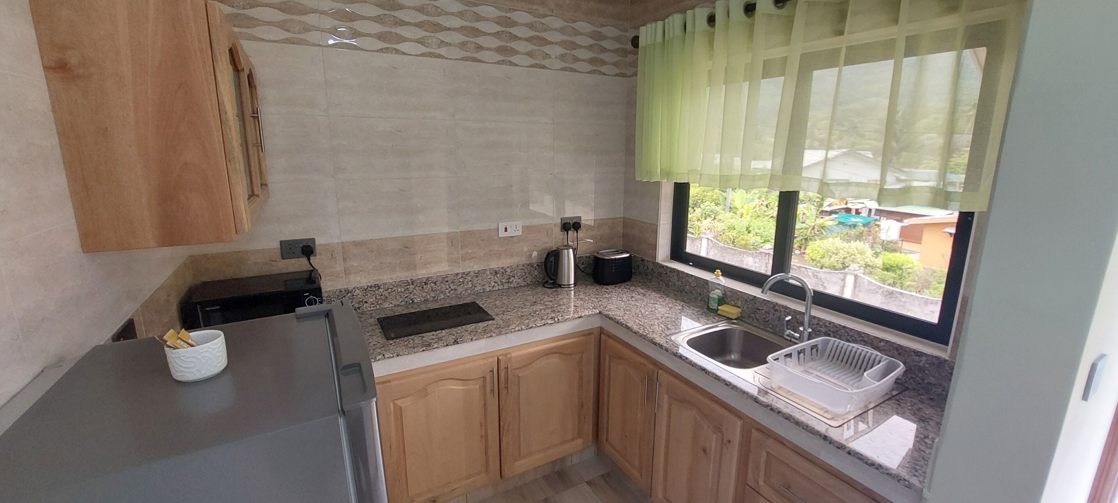 Une cuisine dotée de plans de travail en granit, de meubles en bois, d’un évier en acier inoxydable et d’une fenêtre ornée d’un rideau vert.