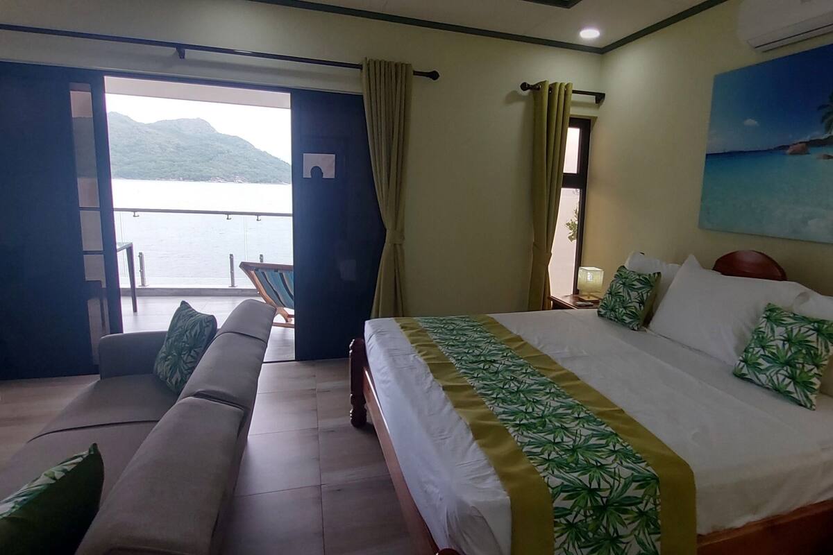 A hotel room with a bed, a sofa, a window with curtains, a painting of a beach, and a view of the sea.
