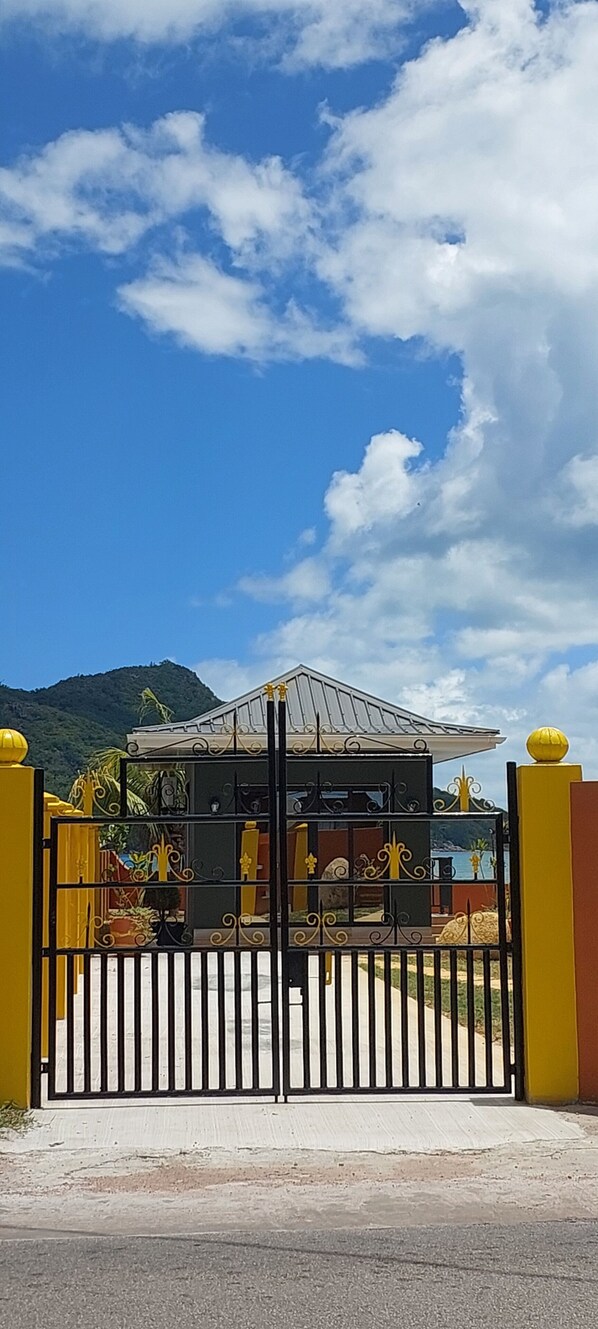 A gated entrance with a metal gate, a covered structure with a roof, and a mountainous background. - Balcons Sur Mer Apartments (Praslin Island)