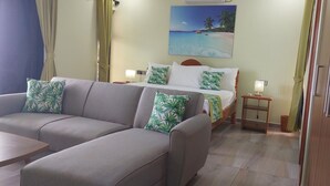 Une chambre d’hôtel avec un canapé, un lit orné d’un tableau représentant une plage tropicale, une table de chevet et une porte en bois.