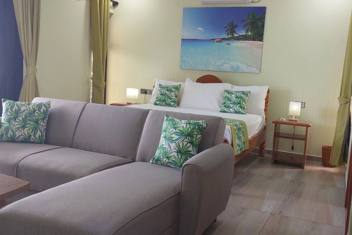 A hotel room with a sofa, a bed with a tropical beach painting, a nightstand, and a wooden door.