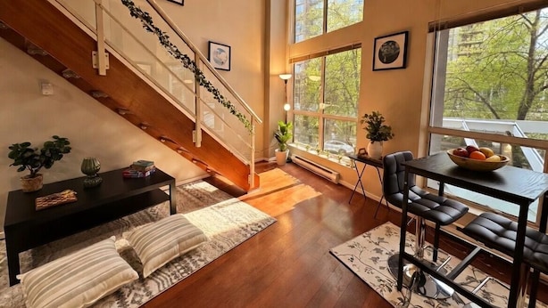 A spacious living area with a wooden staircase, a dining table with chairs, and large windows offering a view of trees. - Urban Loft Perch • City Views • Free Parking (Vancouver)