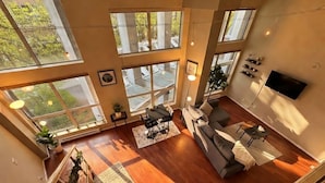 A spacious living room with a large sofa, a coffee table, and a TV mounted on the wall. There are multiple large windows providing natural light and views of the outdoors. - Urban Loft Perch • City Views • Free Parking (Vancouver)