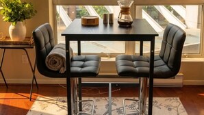 A modern room with a view of buildings and trees, a table with chairs, a potted plant, and a framed picture on the wall. - Urban Loft Perch • City Views • Free Parking (Vancouver)