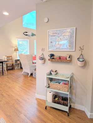 A cozy room with a wooden floor, a wicker chair, a small table, a lamp, a framed map, shelves with baskets, and a cart with storage bins. - Sugar Magnolia 3BR Cottage (Daufuskie Island)