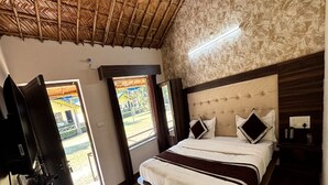 A bedroom with a bed, a TV, a wooden floor, a patterned rug, and a view of the outside. - Nakshatra Resort (Rishikesh)