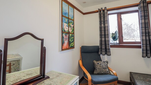 A room with a large mirror, a chair with a cushion, a map on a table, a painting on the wall, and a window with curtains. - Islay Cottage (Kingussie)
