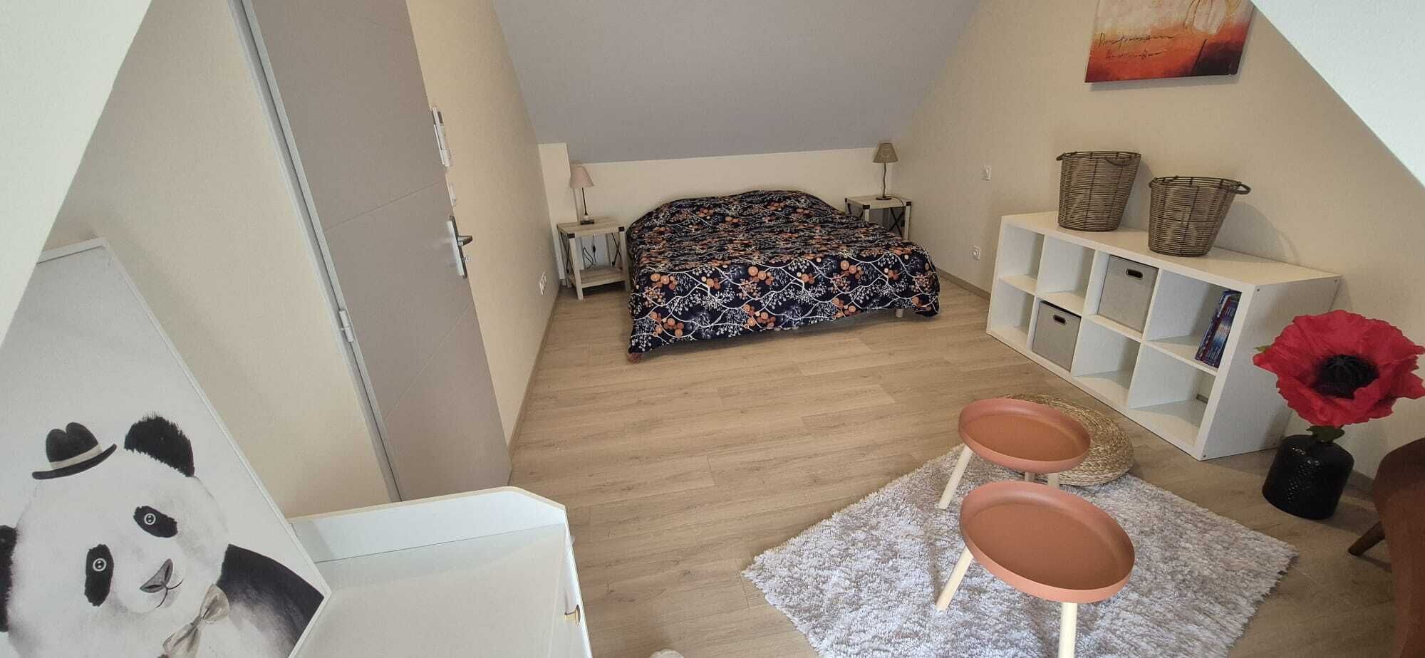 A bedroom with a bed, bedside tables, a nightstand, a bookshelf, a small table with stools, and a decorative vase with a flower.