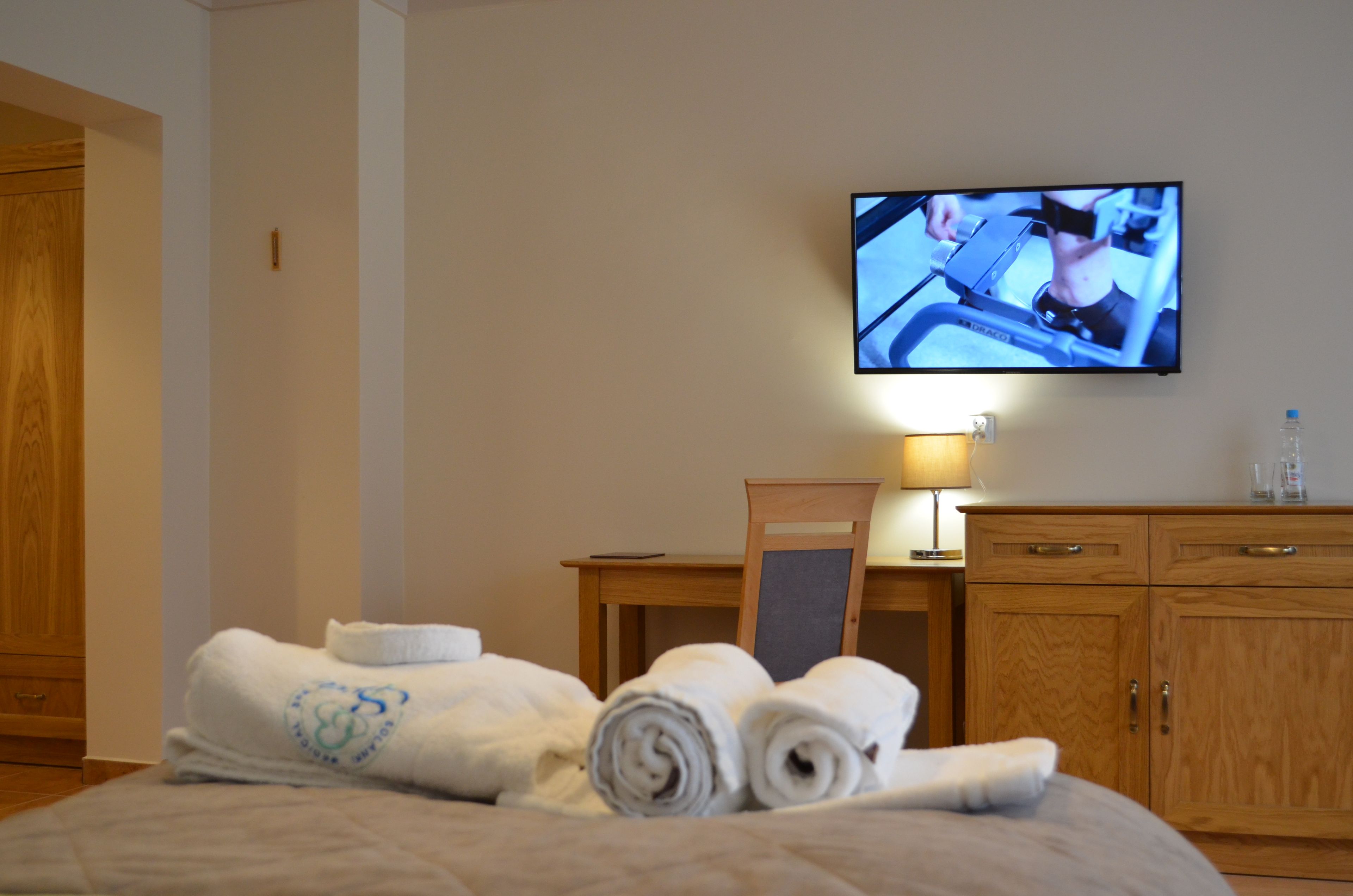 A hotel room with a bed, a television mounted on the wall, a wooden cabinet, and a table with a lamp.